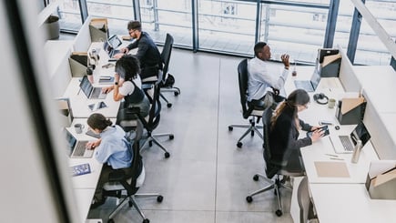 Employees working at desks with laptops and devices in a modern, open-plan office with large windows