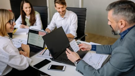 Group of colleagues in a meeting room discussing notes and charts, with laptops and documents on the table