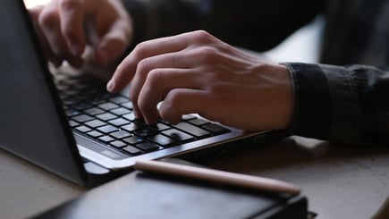 Close-up of hands typing on a laptop keyboard with a notebook and pen nearby