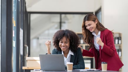 Two businesswomen celebrating success while looking at a laptop in a modern office.