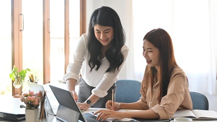 Two women collaborating at a bright office desk with a laptop, notebook, and documents.