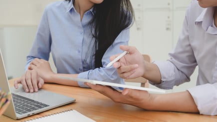 Two people discussing work at a desk, using a tablet and laptop with a notebook nearby.