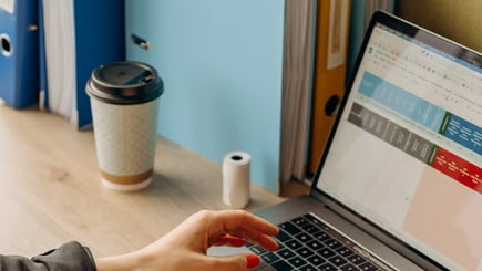 Person working on a laptop with a colorful spreadsheet open, next to office folders and a takeaway coffee cup