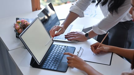 Two people collaborating at a desk with a tablet, laptop, and notepad